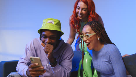 Studio-Shot-Of-Young-Gen-Z-Friends-Sitting-On-Sofa-Sharing-Social-Media-Post-On-Mobile-Phones-3